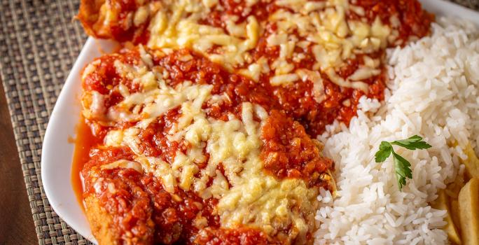 Fotografia de um prato retangular branco com dois filés de frango à parmegiana com queijo derretido por cima, ao lado de um pouco de arroz e batatas fritas.