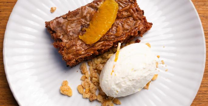 Fotografia de um prato branco raso com um pedaço de bolo de chocolate cremoso com crocante de nozes, e por cima, chantilly de laranja. O prato está sobre uma bancada de madeira clara.