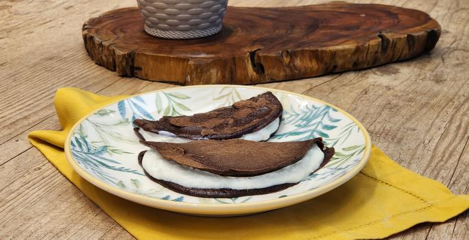 Fotografia em tons de marrom com um prato redondo ao centro. Em cima do prato existe duas panquecas de chocolate com aveia recheadas com um creme de iogurte.