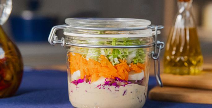 Fotografia em tons de azul com um pote de vidro ao centro. Dentro do pote existe uma receita de salada de repolho roxo feita com repolho, cenoura, palmito e molho rosé