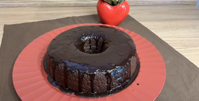 Fotografia de um bolo de chocolate e café com calda de chocolate sobre um apoio grande vermelho. O apoio está por cima de um pano marrom de tom escuro.