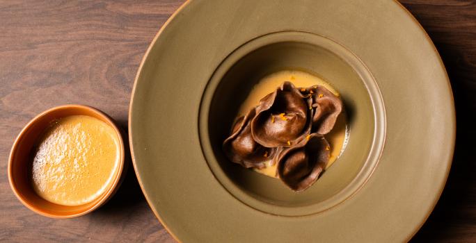 Fotografia vista de cima de um capeletti de cacau e café, recheado com creme de banana, por cima de um creme anglaise de café. A receita está em um prato fundo de bordas grossas e cor verde, ao lado tem um recipiente fundo pequeno com mais molho.