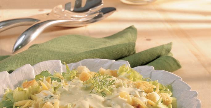 Fotografia em tons de verde em uma bancada de madeira clara, um pano verde, um pegador de salada e um prato redondo branco fundo com a salada de alface, queijo, torradinhas e iogurte dentro.