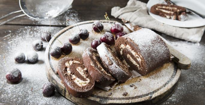 Fotografia de um rocambole de pão-de-ló de chocolate, recheado com Leite MOÇA, coco e castanha-de-caju. Por cima tem um povilhado de açúcar de confeiteiro