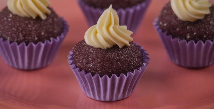 Fotografia de quatro forminhas de cor roxa com um brigadeiro de açaí, e por cima de cada um tem uma cobertura de chocolate confeitado. As unidades de brigadeiro estão sobre um apoio de cor rosa.