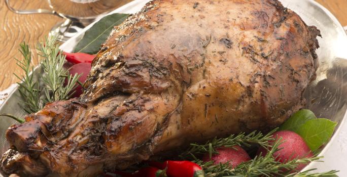 Fotografia em tons de marrom e verde, com travessa com duas alças contendo pernil assado guarnecido com ervas e pimenta vermelha, sobre dois guardanapos com um garfo, acima pratos empilhados, potinho e molheira com molhos, tudo sobre bancada de madeira.