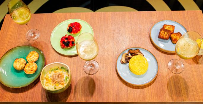 Fotografia de uma mesa de madeira com um brunch com cuscuz, quiche de alho poró, cheesecake, salada de frutas e suco de abacaxi com hortelã. As comidas estão em pratos coloridos, e o suco estão em taças, na imagem são três.