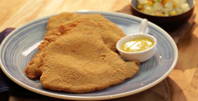 Fotografia em tons de marrom e azul de uma bancada de madeira clara, sobre ela um prato azul com dois bifes á milanesa e um mini recipiente refratário com molho.  Ao fundo um recipiente pequeno azul e redondo com vinagrete.