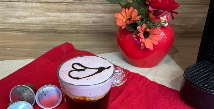 Fotografia em tons de vermelho em uma bancada de madeira, um pano vermelho e uma xícara com café e espuma rosa decorado com um coração com calda de chocolate ao centro. Ao fundo, um vaso vermelho com flores e em formato de coração.