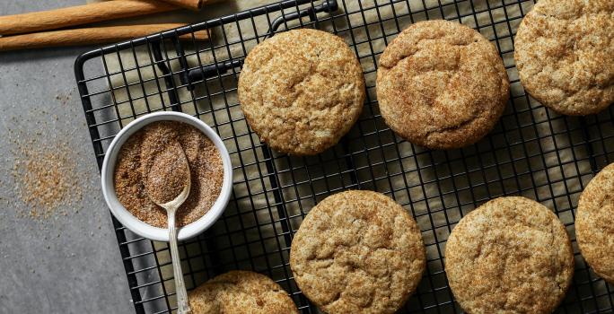 Fotografia vista de cima de sete cookies com açúcar e canela sobre uma grade preta. Ao lado dos cookies e por cima da grade, tem um pequeno pote de vidro branco com açúcar e canela, e uma colher de chá apoiada sobre ele.