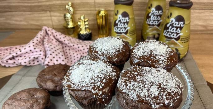 Fotografia em tons de marrom e dourado com um recipiente de vidro ao centro. Em cima do recipiente existe vários muffins cobertos com chocolate e coco ralado. Ao fundo existe três garrafas de PANTÁSTICA, uma mistura pronta para panquecas.