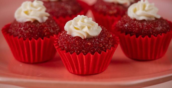 Fotografia de cinco unidades forminhas de cor vermelha, um junto da outra, e dentro de cada forminha tem um brigadeiro de red velvet, passado no açúcar cristal com uma decoração de cream cheese. Os brigadeiros estão sobre um apoio decorativo rosa alto.