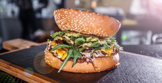 Fotografia de um hamburguer de carne e champignon temperado e servido com rúcula. A fatia do pão na parte de cima, está ao contrário, e o lanhe está sobre uma tábua de madeira com fundo preto.