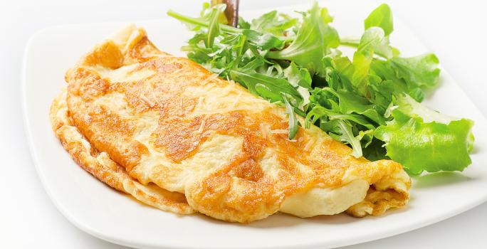 Fotografia de um omelete com crosta de queijo crocante dobrado ao meio, ao lado de uma salada de folhas, sobre um prato de vidro branco. O prato está sobre uma mesa branca.