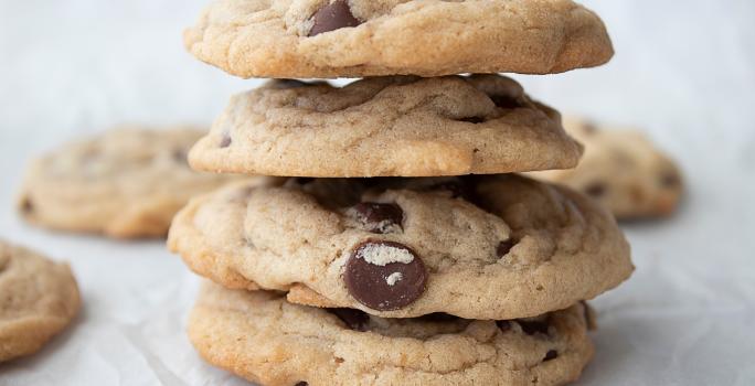 Fotografia de quatro cookies de leite condensado e baunilha empilhados, todos têm gostas de chocolate. Os cookies estão sobre uma mesa branca, ao fundo, tem mais unidades deles, espalhados.