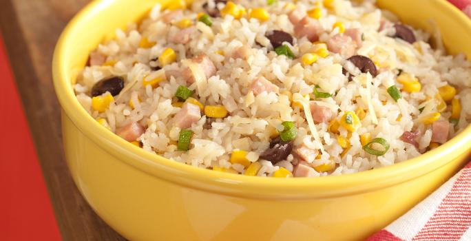 fotografia em tons de vermelho, marrom e amarelo de uma bancada marrom vista de frente, acima da bancada contém um recipiente redondo amarelo com arroz peito de peru e milho. Ao lado um pano xadrez em tons de vermelho e branco.