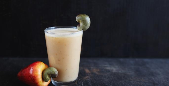 Fotografia de um copo de vidro com refresco de Neston com suco de caju, ao lado de um caju in natura sobre uma mesa escura e um fundo preto.