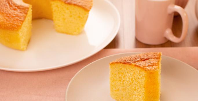 Fotografia de um prato raso branco grande com um bolo quase inteiro de fubá, na frente tem outro prato, menor e de tom mais escuro, com uma fatia do bolo de fubá. Um pouco mais ao fundo tem uma xícara rosa com café.