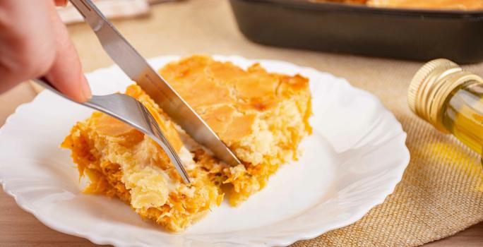 Fotografia de uma fatia de trota de frango em um prato raso de vidro branco. Tem uma mão masculina cortando um pedaço com garfo e faca. No fundo tem a uma forma preta com a torta assada.