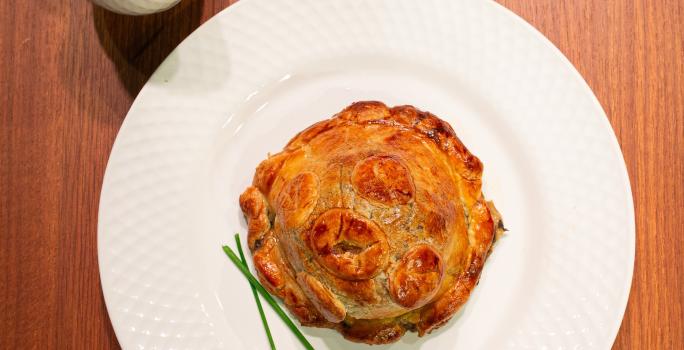 Fotografia de uma receita de pithivier paraense com duxelles de ora-pro-nóbis sobre um prato raso branco, o qual está sobre uma bancada de madeira. Ao lado do prato tem um recipiente fundo e pequeno com molho.