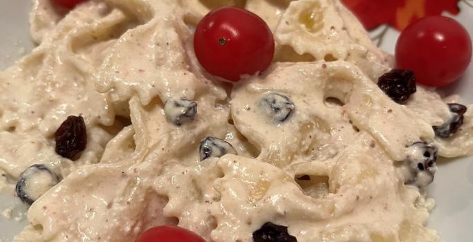 Fotografia de uma masssa farfalle cremosa com uvas passa e tomates cereja dentro de um prato branco de vidro.