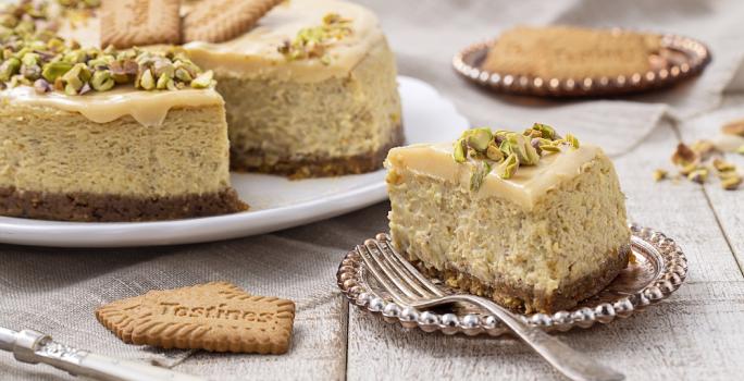 Foto da receita de Cheesecake de Pistache. Observa-se a torta com um pedaço cortado em um prato de sobremesa, decorado com pistaches e uma calda na cor branca.