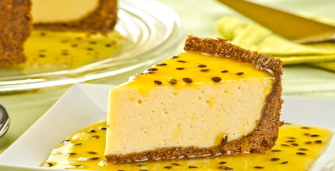 Fotografia de uma fatia grande de torta de maracujá com base de biscoitos e calda da fruta sobre e por volta da fatia, que está em um prato quadrado branco. No fundo da foto tem a torta inteira com mais calda de maracujá.