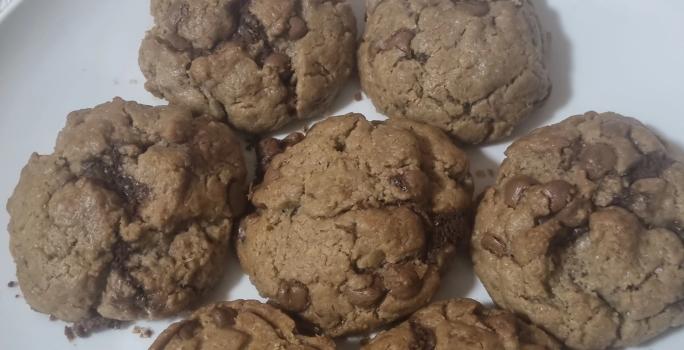 Fotografia de sete cookies grandes com gotas de chocolate sobre um prato grande e raso, de cor branco.