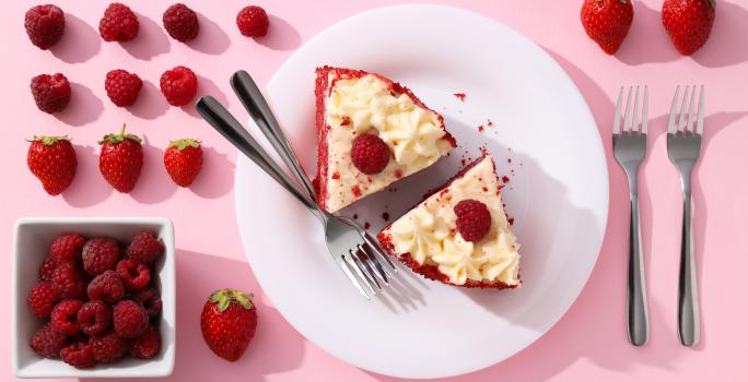 Fotografia vista de cima de um prato raso grande branco de vidro com duas fatias de torta red velvet, com dois garfos juntos ao lado. Ao redor do prato tem mais dois garfos e muitas frutas vermelhas.