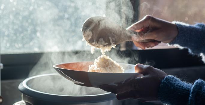 Fotografia de uma mão feminina colocando com uma colher de pau um arroz cremoso ao champanhe em um prato fundo. A imagem está contra a luz do sol, e se caracteriza pelo vapor saindo da panela.