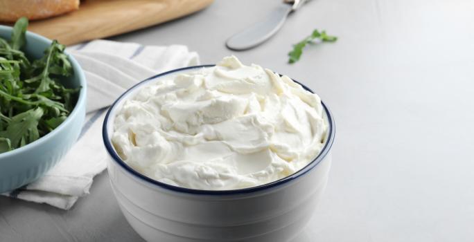 Fotografia em tons de branco com um pote branco ao centro. Dentro do pote existe uma receita de cream cheese caseiro, um creme branco feito com vinagre, leite e creme de leite.