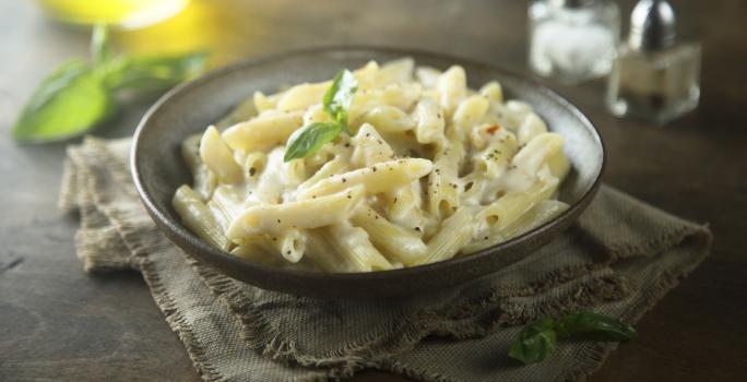 Fotografia em tons de cinza de uma bancada de madeira, ao centro um paninho cinza amarronzado com um prato redondo sobre ele com o macarrão pene com molho branco.