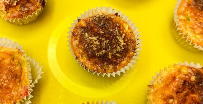 Fotografia em tons de amarelo com vários muffins de omelete ao centro. Em cima de cada muffin existe uma camada de queijo parmesão