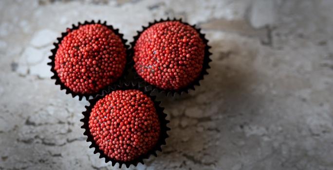 Fotografia em tons de vermelho, em uma bancada de madeira de cor cinza. Ao centro, 3 formilhas de brigadeiro contendo os brigadeiros.