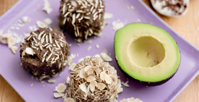 Fotografia em tons de roxo com um prato retangular roxo ao centro. Em cima do prato existe 3 brigadeiros de cor marrom e ao lado existe metade de um abacate. Todos os brigadeiros estão polvilhados com coco ralado