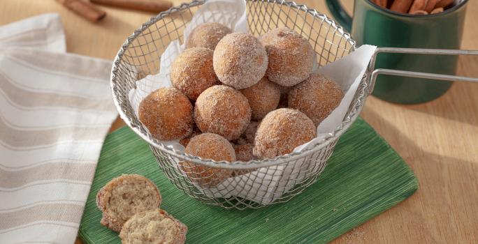 Foto da receita de bolinho de chuva sem glúten. Observa-se uma peneira com os bolinhos dentro, polvilhados de açúcar e canela, sobre um guardanapo de pano verde