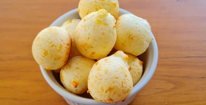 Fotografia de um ramekin branco com vários pães de queijo. O recipiente está sobre uma mesa de madeira.