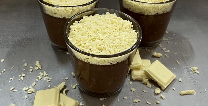 Foto da receita de trufa cremosa servida em três porções em copos de vidro baixos sobre uma mesa acinzentada com pedaços e raspas de chocolate branco rodeando os copos como decoração