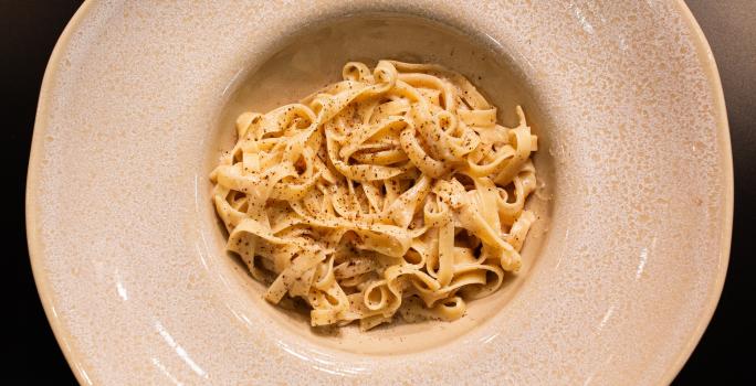 Fotografia em tons de cinza e preto com um prato cinza ao centro. Em cima do prato existe uma receita de cacio e pepe feita com macarrão tagliatelle e finalizado com pimenta preta moída