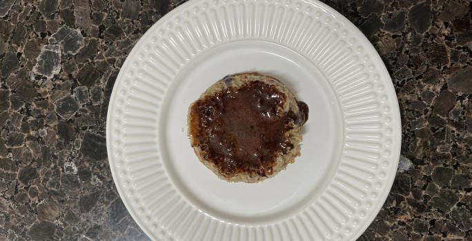 Fotografia vista de cima de um prato raso de vidro branco com um bolinho de micro-ondas com uma calda de chocolate feita de whey. O prato está sobre uma bancada escura.