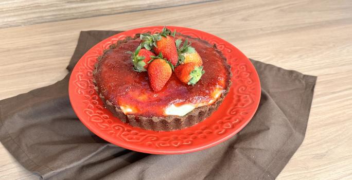 Fotografia em tons de vermelho com um prato vermelho ao centro. Em cima do prato existe um cheesecake de Galak com queijo gorgonzola, cobertura de morango e morangos inteiros