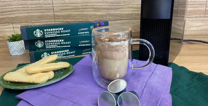 Fotografia em tons de verde e roxo em uma bancada de madeira, um paninho verde e roxo ao centro com uma xícara de vidro e o café tiramisu dentro. Ao fundo, uma máquina nespresso.