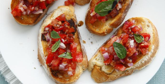 Fotografia vista de cima de cinco fatias de pão italiano torrado com uma bruschetta por cima. Os aperitivos estão sobre um prato de vidro branco, que está apoiado em um pano de mesa de cor azul.