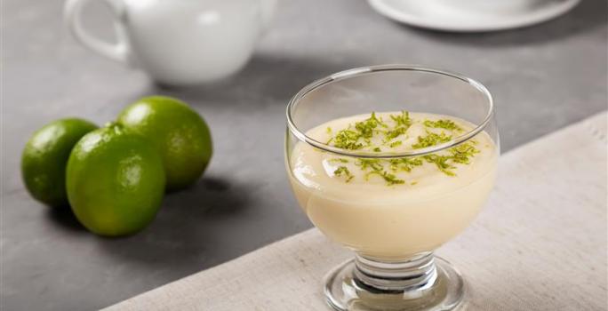 Fotografia em tons de branco com uma taça de sobremesa de vidro ao centro dentro da taça existe uma receita de mousse de limão coberta com raspas de limão.