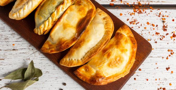 Fotografia vista de cima de cinco pasteis assados na air fryer sobre uma tábua retangular de madeira pequena, a qual está sobre uma mesa de madeira branca.