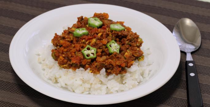 Fotografia em tons de marrom com um prato branco fundo ao centro. Dentro do prato existe uma porção de arroz coberta com carne moída e pedaços de quiabo. Ao lado existe uma colher.