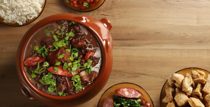 Fotografia vista de cima de uma feijoada completa, com cebolinha por cima, em um recipiente grande clássico de vidro marrom. Ao redor tem acompanhamentos de arroz, torresmo, vingrete e couve, todos em um recipiente de vidro, sobre uma mesa de madeira