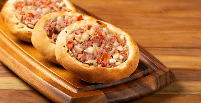 Fotografia de três esfihas de carne, uma apoiada na outra, sobre um apoio de madeira retangular, o qual está apoiado em uma mesa de madeira.