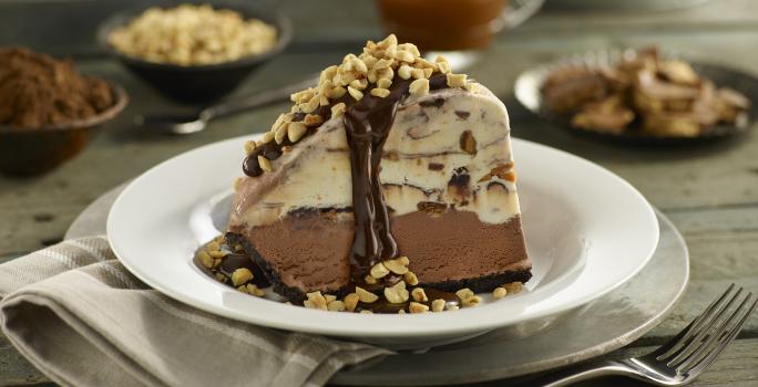 Fotografia de uma fatia de torta Alaska feita de sorvete de café com uma calda de chocolate e amendoins triturados. A torta está em um prato fundo branco.