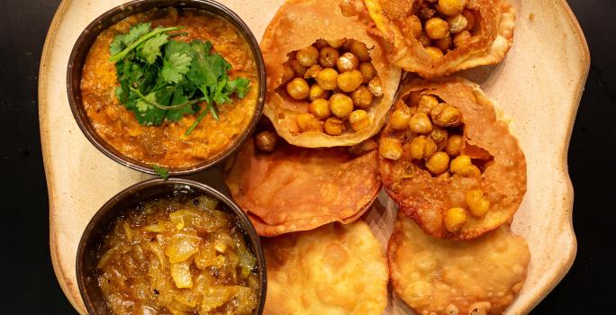 Fotografia vista de cima de seis unidades de pani puri com grão de bico Crocante, e ao lado tem dois recipientes pequenos, um com chutney de cebola e o outro com dahl de lentilha. A receita está em um prato de vidro branco, sobre uma mesa marrom.